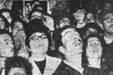 A crowd of men and women looking upward at night, faces lit